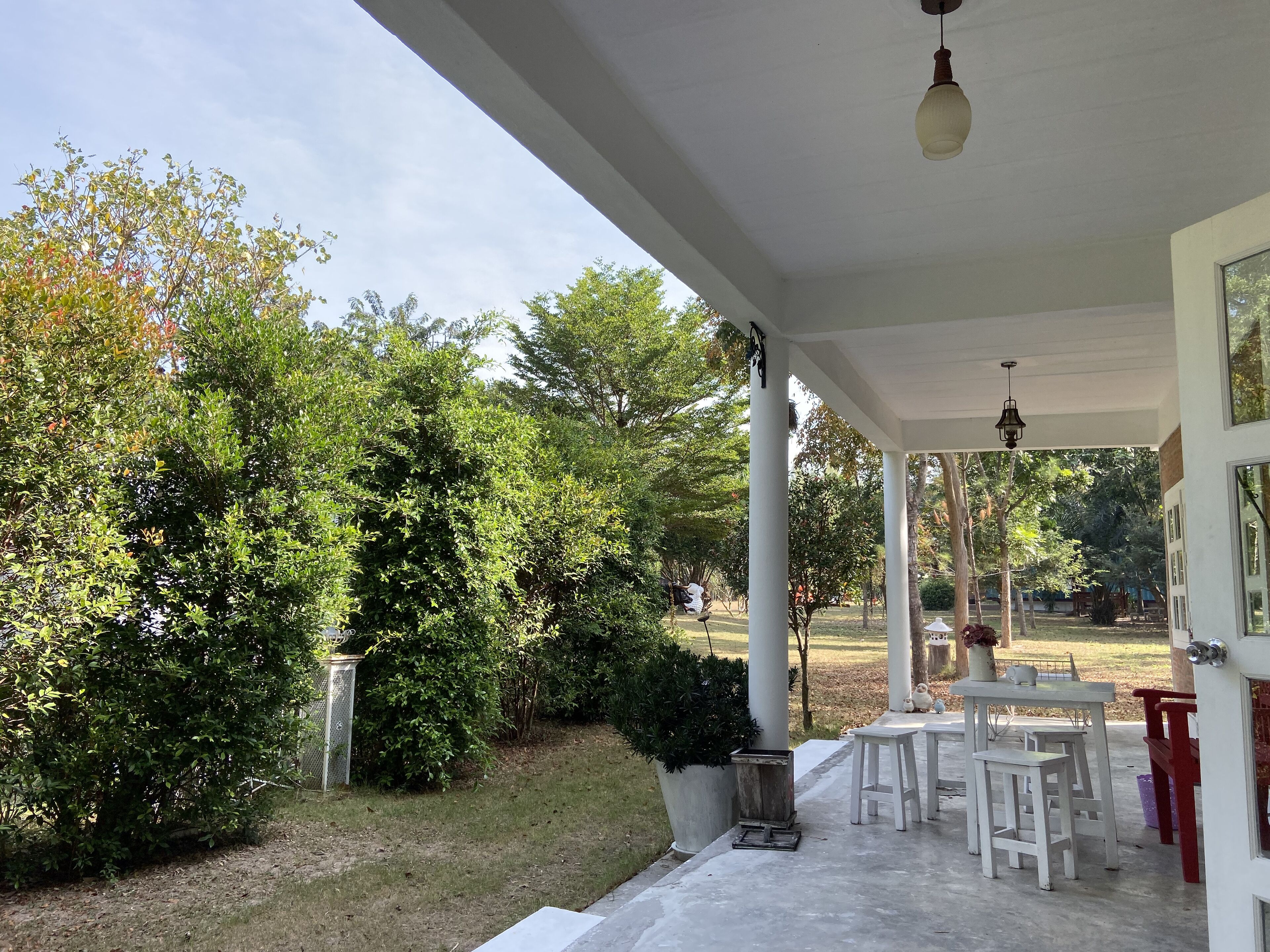A covered patio area with white columns, a table with chairs, and a view of a garden with various trees and shrubs.