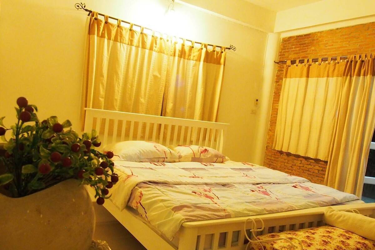 A bedroom with a white bed, yellow curtains, a brick wall, and a plant with red berries.