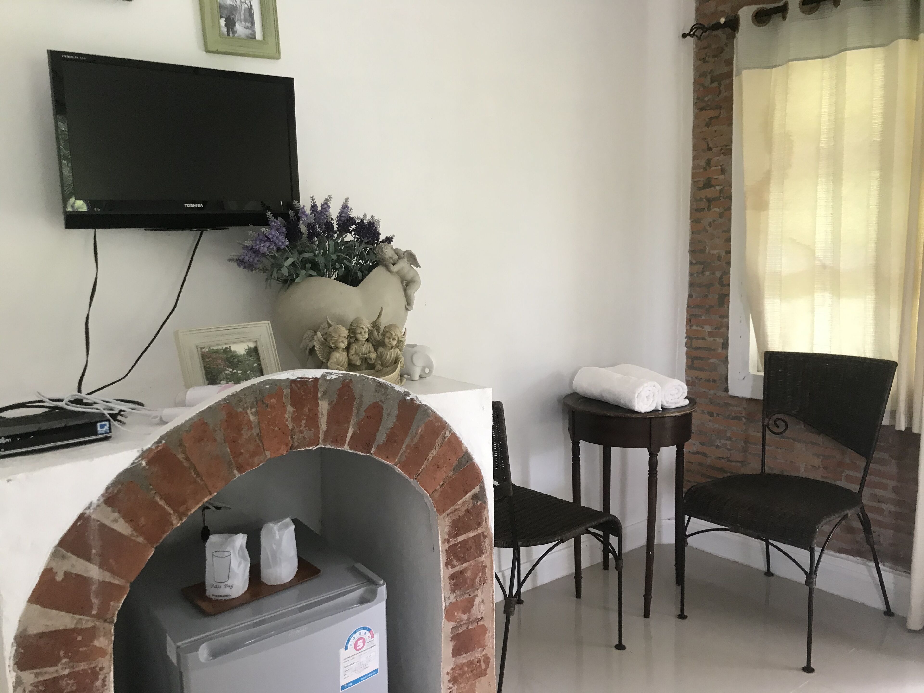 A room with a television mounted on the wall, a heart-shaped planter with flowers, a small table with towels, a brick oven, and two chairs.