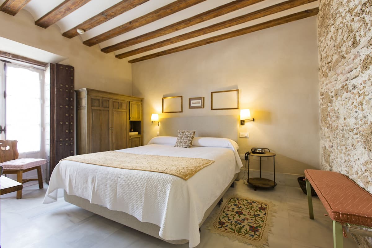 A bedroom with a large bed, wooden beams on the ceiling, a small table, a bench, and a wall with a textured pattern.