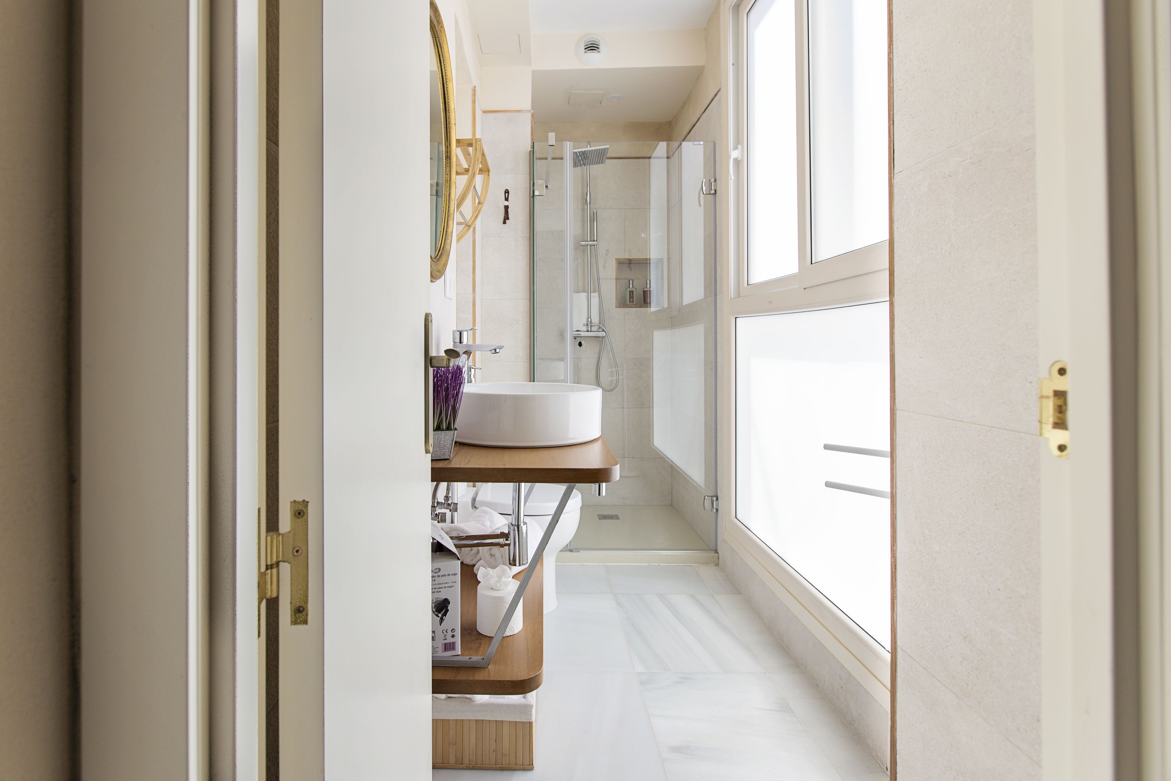 A narrow, well-lit bathroom with a large window, a sink, and a shower.
