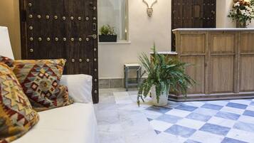 Una habitación con piso a cuadros, una puerta de madera con remaches metálicos, un sofá blanco con cojines estampados, una mesita y una planta en maceta.