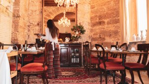A woman standing in a dining area with a brick wall, wooden furniture, and a chandelier. - Hotel Casa de las Cuatro Torres (Cádiz)