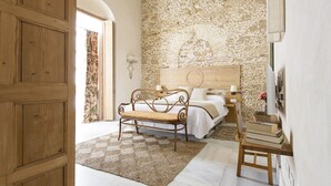 A bedroom with a stone wall, a wooden bench, a bed with white linens, and a bedside table with books. - Hotel Casa de las Cuatro Torres (Cádiz)