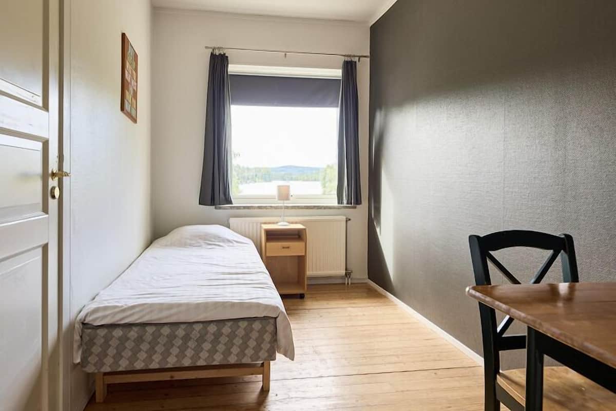 A small, single-bed room with a wooden floor, a window with curtains, a bedside table, and a chair.
