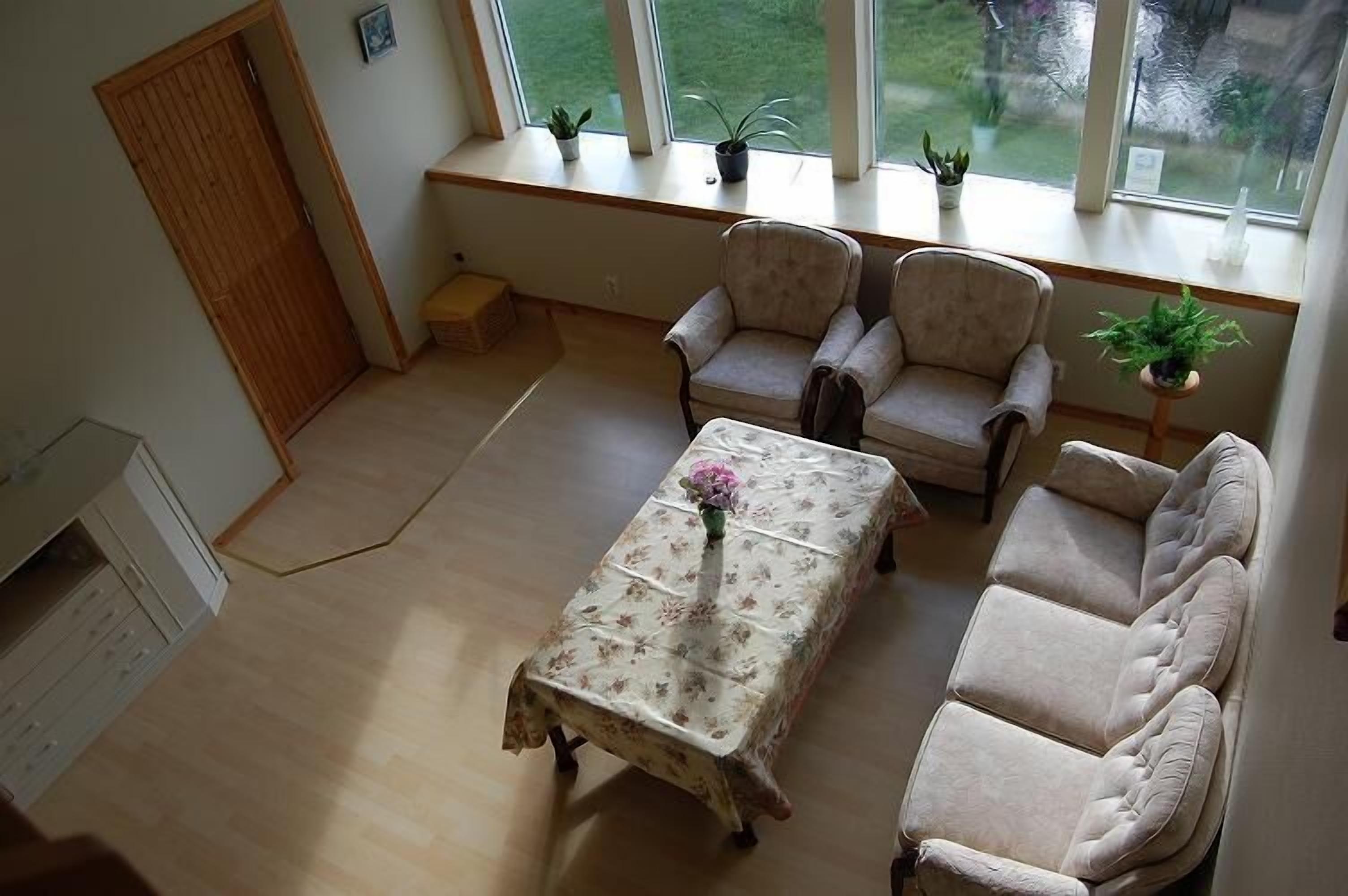 A living room with a sofa, two armchairs, a coffee table with a floral tablecloth, and a window with plants.