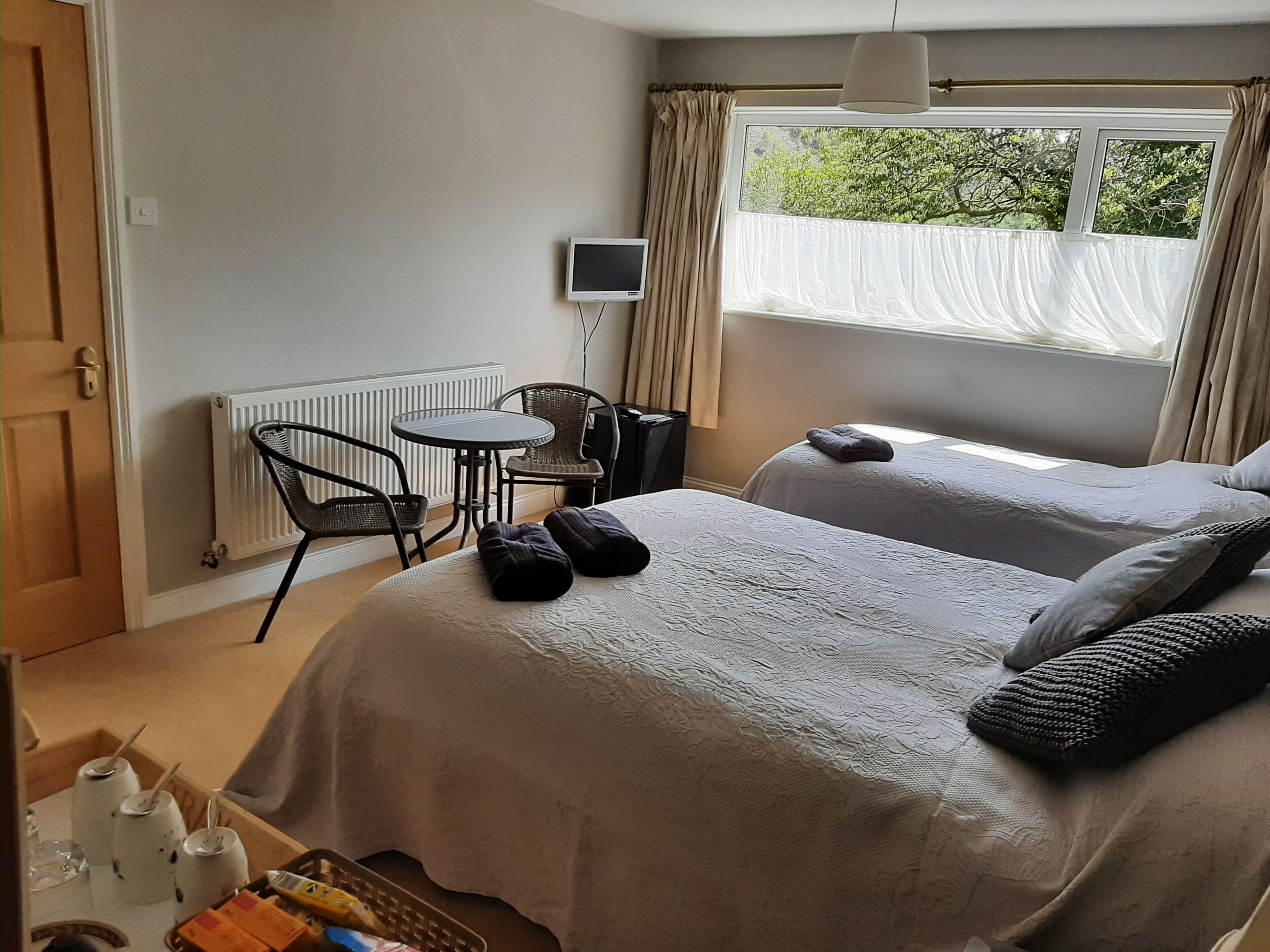 A hotel room with two beds, a small table with chairs, a radiator, and a window with curtains.