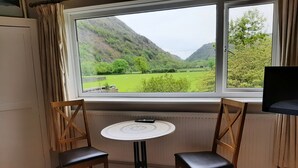 Quarto com vista para um vale verde e montanhas através de uma janela. Há uma mesa redonda com duas cadeiras e um controle remoto sobre ela.