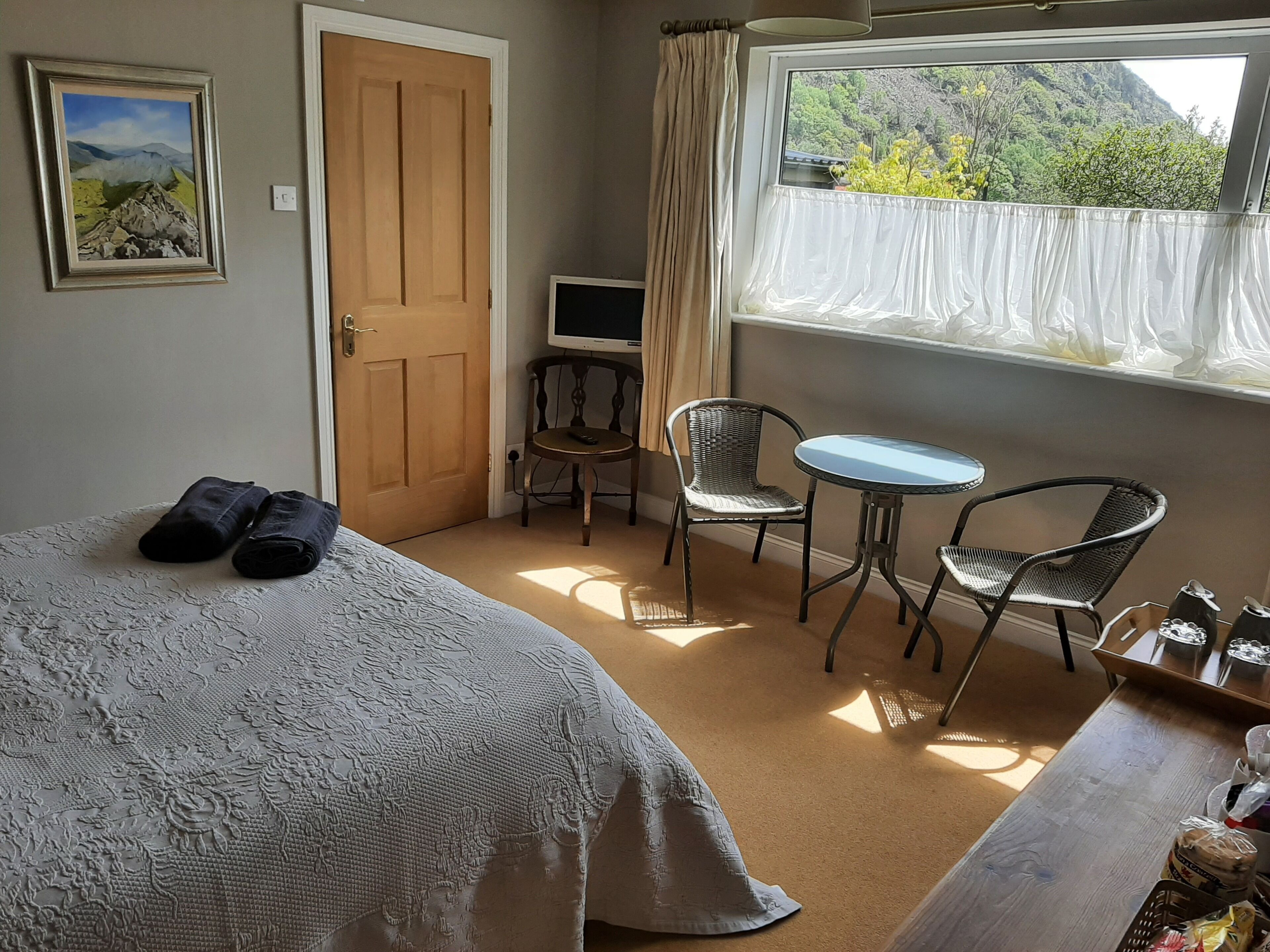 Quarto com cama, escrivaninha, duas cadeiras, televisão e janela com vista para área verde.