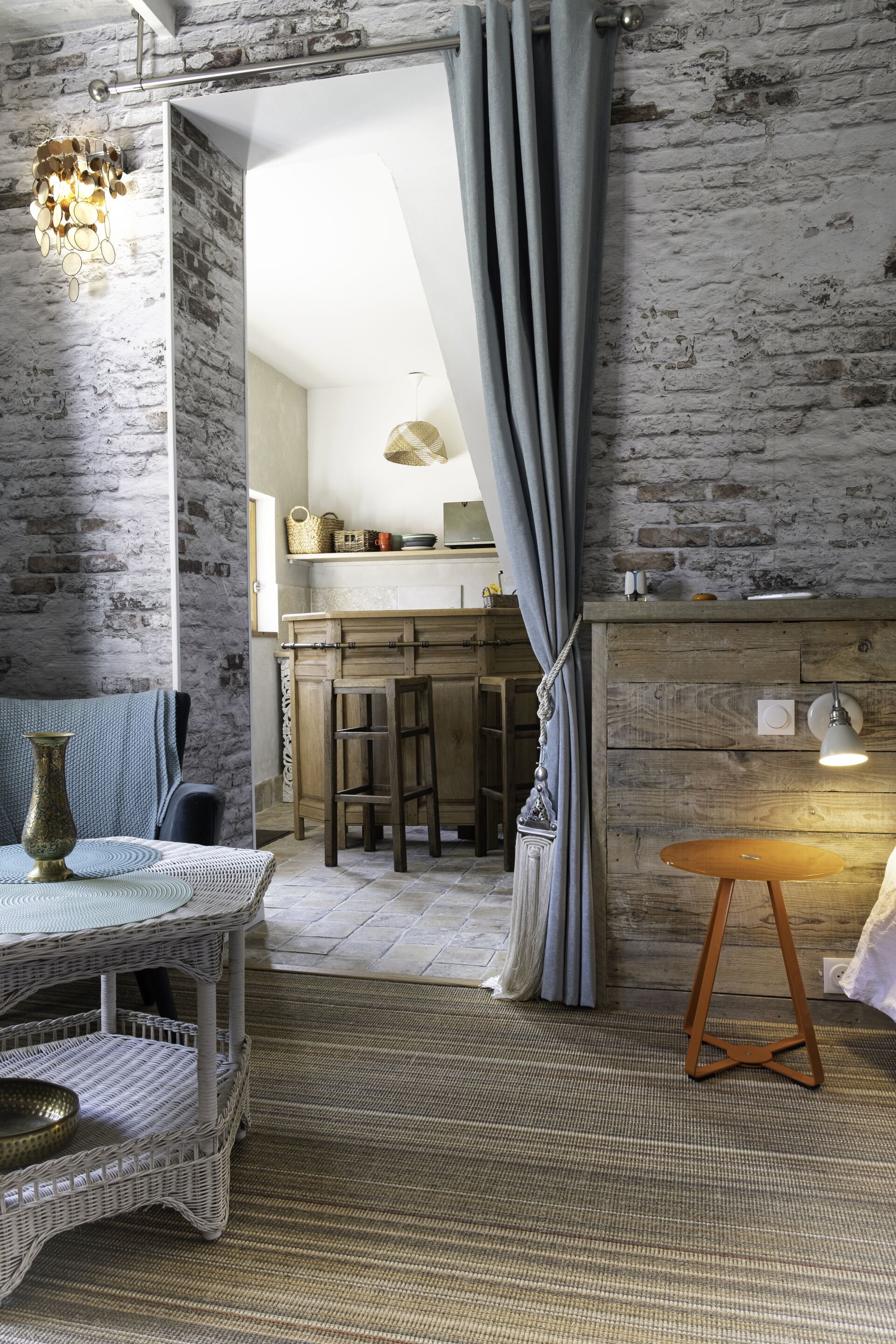 A rustic interior with exposed brick walls, a dining area with a wicker table and chairs, and a kitchen visible through an open doorway.