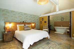 Quarto com cama grande, cabeceira de madeira, luminárias de cabeceira, mesa de cabeceira, tapete estampado e banheiro com banheira e toucador de madeira.