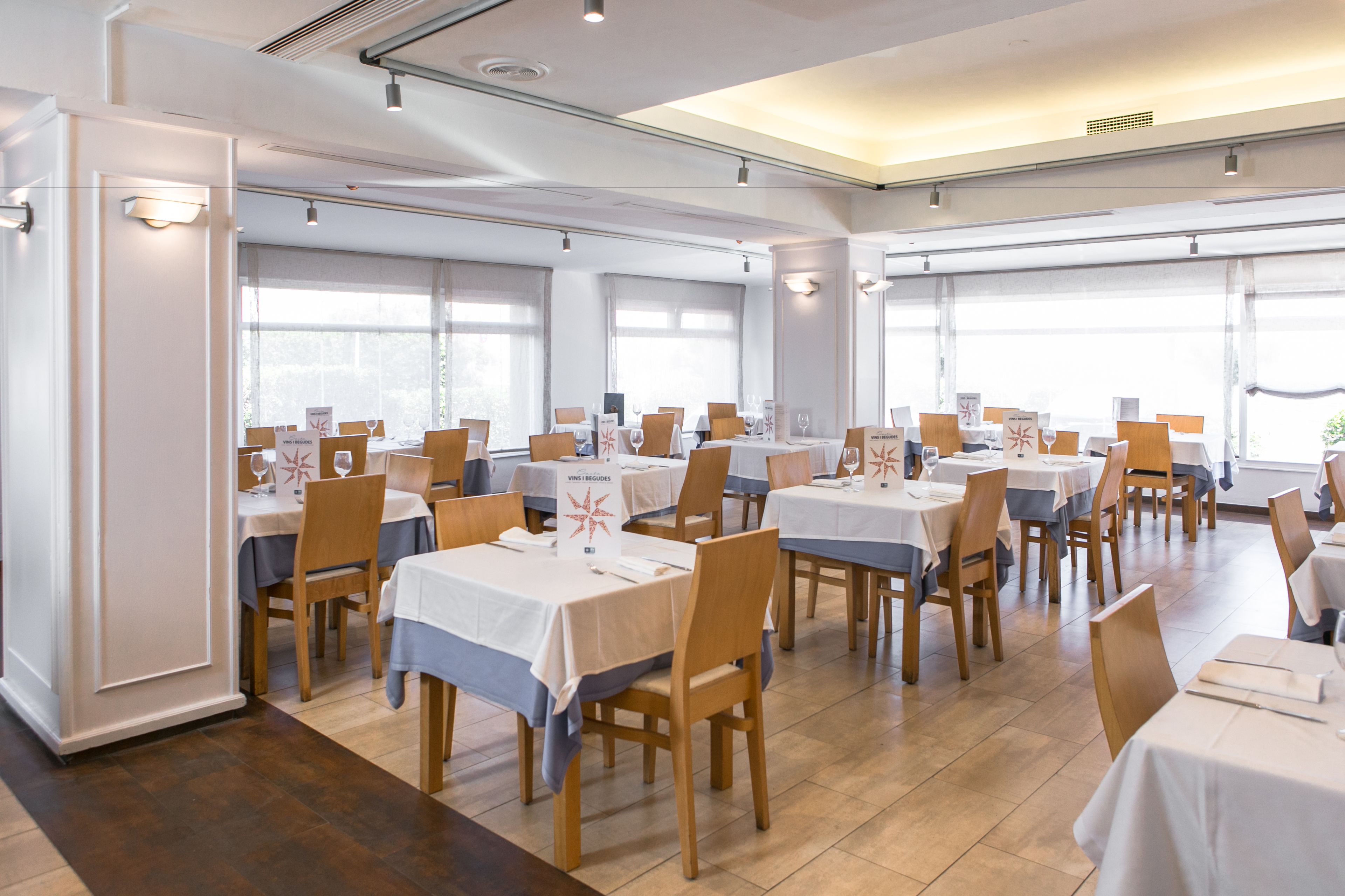 Un comedor con varias mesas y sillas dispuestas en filas, con amplios ventanales que dejan entrar luz natural.