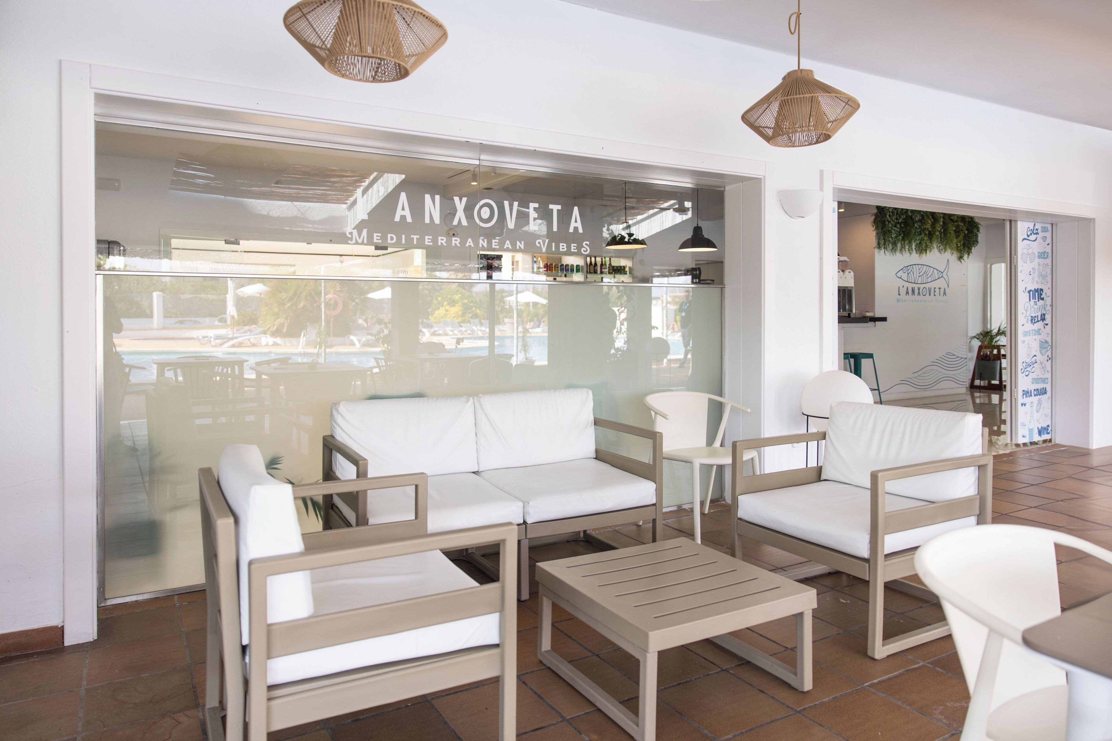 A modern interior with a white sofa, wooden chairs, a small table, and a glass wall with a sign reading 'ANXOVETA Mediterranean Vibes'.