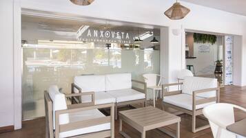 A modern interior with a white sofa, wooden chairs, a small table, and a glass wall with a sign reading 'ANXOVETA Mediterranean Vibes'.