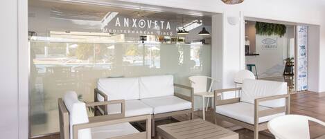 A modern interior with a white sofa, wooden chairs, a small table, and a glass wall with a sign reading 'ANXOVETA Mediterranean Vibes'.