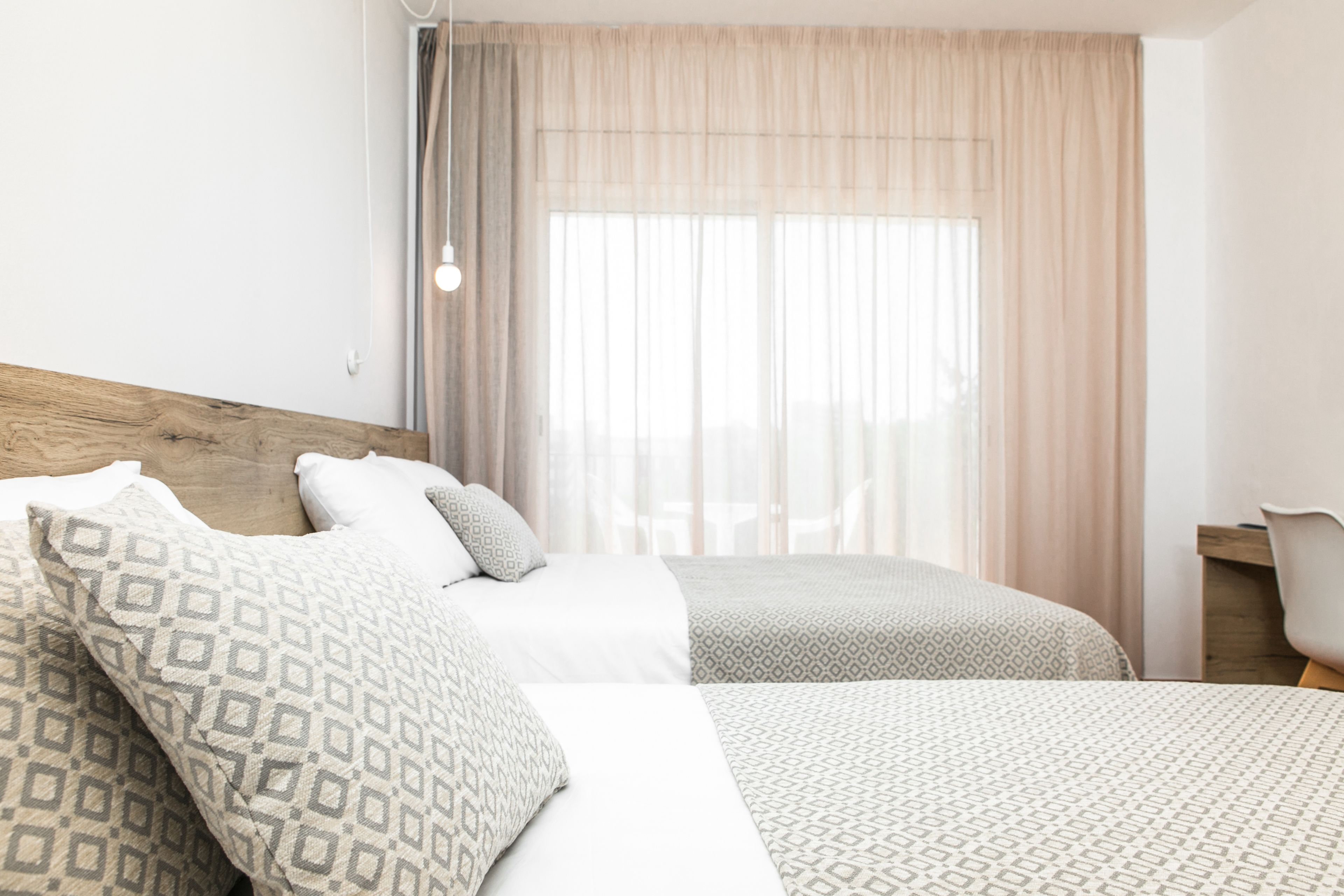 Habitación de hotel con dos camas, cabecera de madera, un escritorio y un ventanal con cortinas traslúcidas.