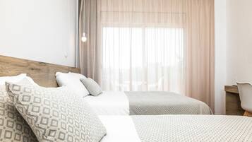 A hotel room with two beds, a wooden headboard, a desk, and a large window with sheer curtains.
