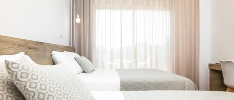 Habitación de hotel con dos camas, cabecera de madera, un escritorio y un ventanal con cortinas traslúcidas.