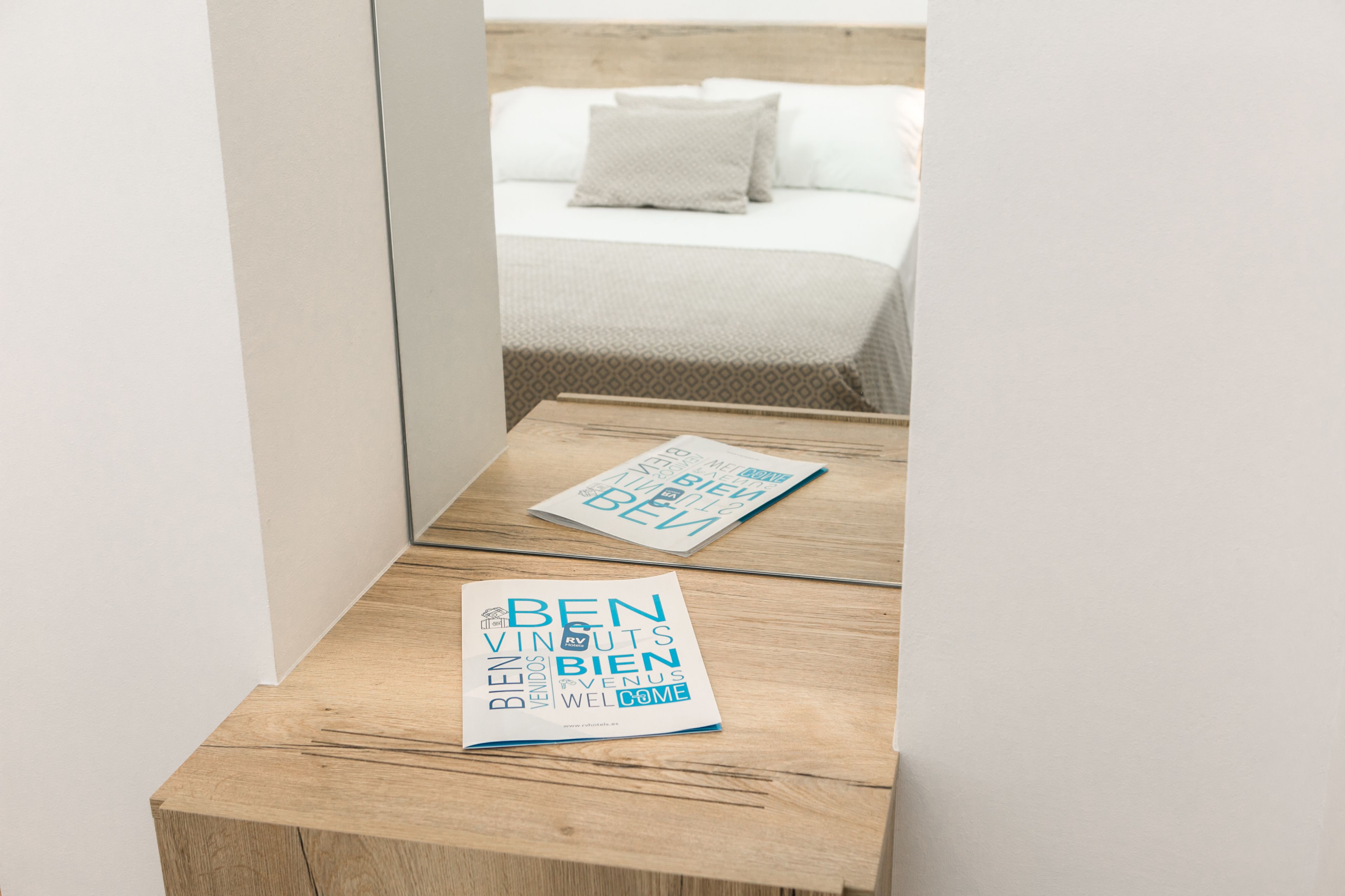 Una mesita de noche de madera con una tarjeta de bienvenida y un espejo que refleja una cama con cabecera blanca y almohadas.