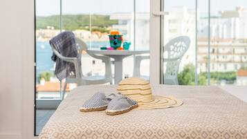 Uma varanda com vista para o mar, uma cadeira branca, uma mesa pequena com brinquedos, e um chapéu de palha e chinelos no chão.