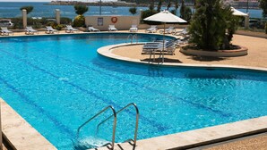 A swimming pool with sun loungers and a lifebuoy, surrounded by a pebble beach and a view of the sea.