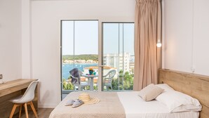 A hotel room with a bed, a desk, and a chair. There is a view of the sea and buildings through large windows.