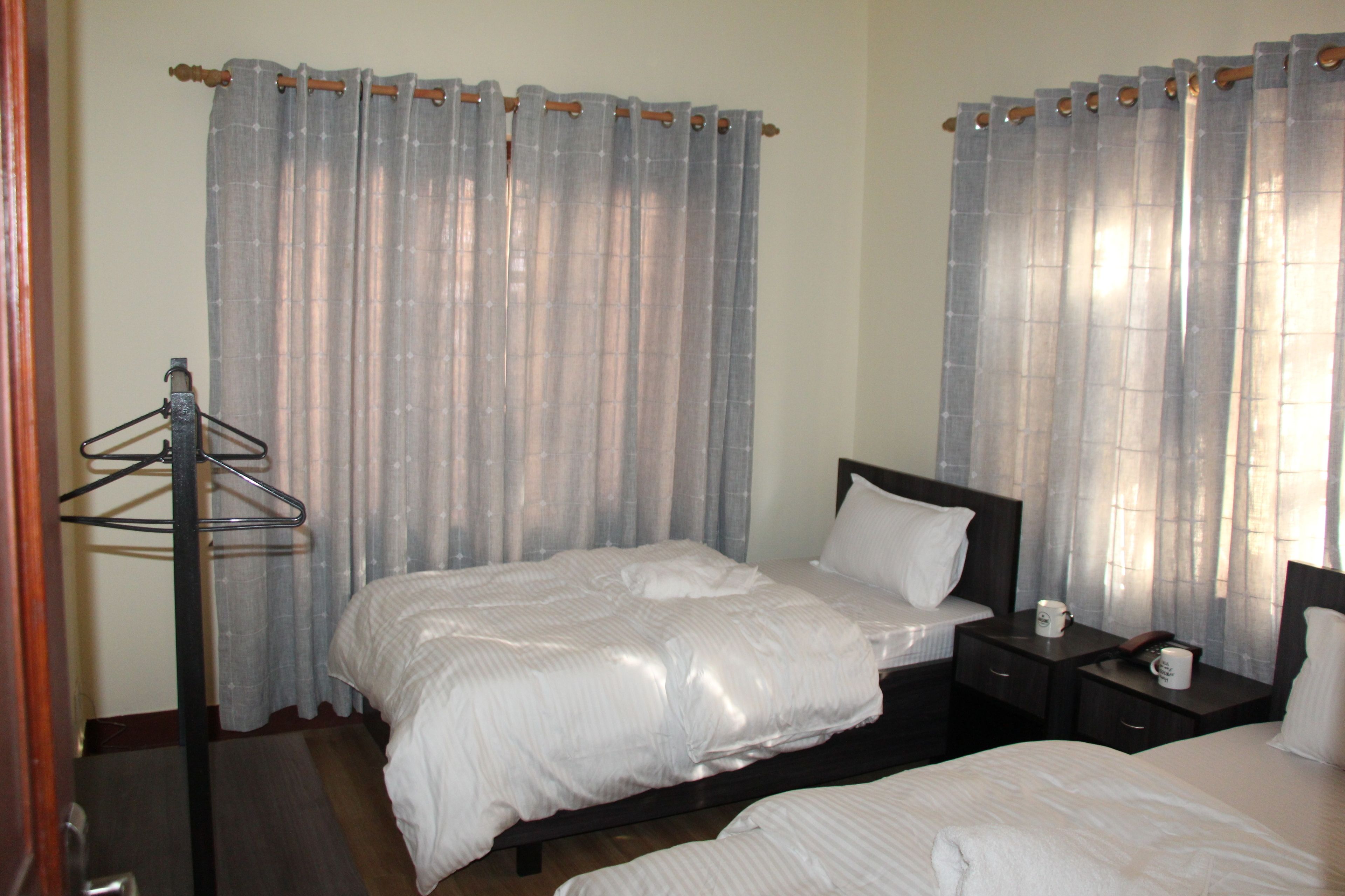 A hotel room with two beds, a wooden nightstand, a black coat rack, and sheer curtains.
