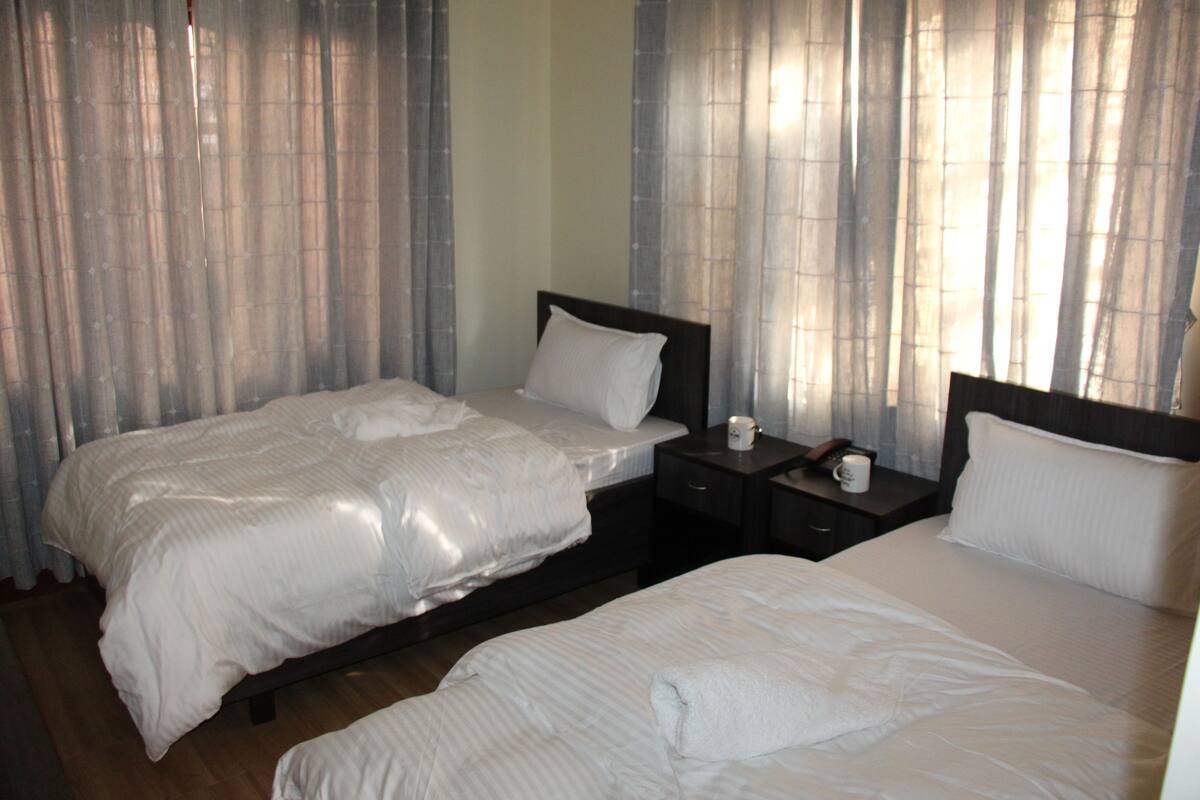 A hotel room with two beds, a bedside table with a lamp, and a window with curtains.