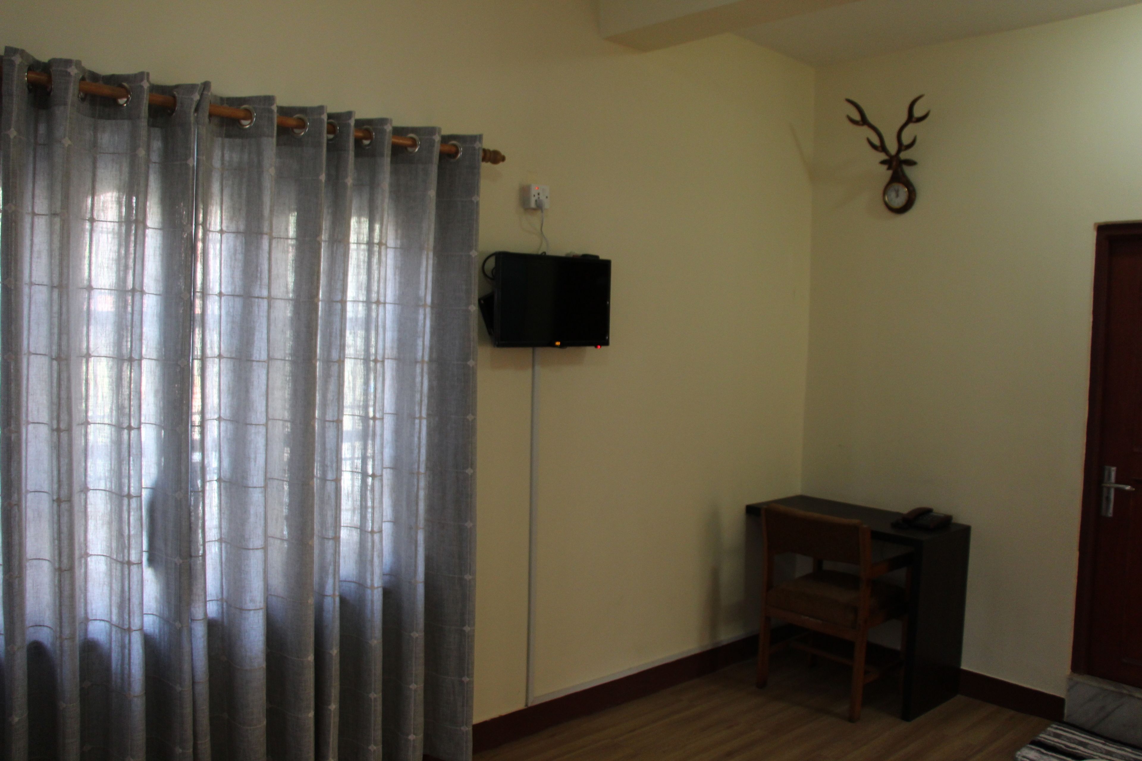 A room with a wooden floor, a table with a chair, a curtain, a mounted clock, and a door.