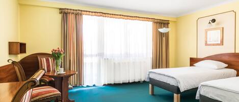 A hotel room with two single beds, a wooden chair, a small table with a vase of flowers, and a large window with curtains.