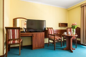 Habitación de hotel con un escritorio de madera, un televisor de pantalla plana, dos sillas a rayas, una mesita con un jarrón de flores y una alfombra azul.