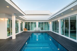 Une piscine intérieure moderne avec une terrasse en bois, de grandes fenêtres et un plafond blanc.