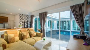A modern living room with a sofa, a coffee table, and a desk with a computer. There is a large window with curtains and a view of a pool and outdoor seating. - Baan Marwin Hua Hin (Hua Hin)