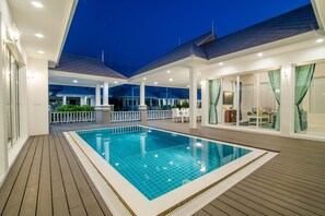 Une piscine avec une terrasse en bois, entourée d’un espace couvert comprenant des sièges et offrant une vue sur le ciel.