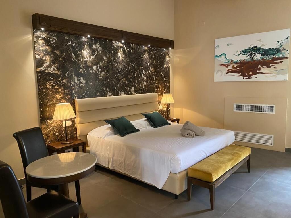 Habitación de hotel con una cama grande, dos mesitas de noche con lámparas, una mesa redonda, un banco y una pared con un diseño similar al mármol y un cuadro.
