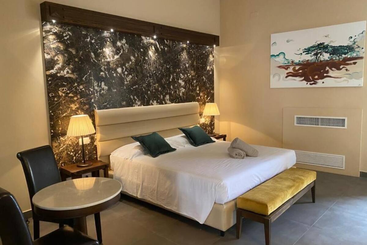 A hotel room with a large bed, two bedside tables with lamps, a round table, a bench, and a wall with a marble-like design and a painting.