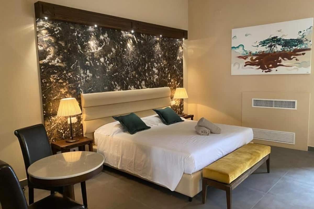 Habitación de hotel con una cama grande, dos mesitas de noche con lámparas, una mesa redonda, un banco y una pared con un diseño similar al mármol y un cuadro.