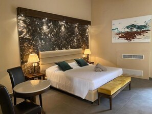 A hotel room with a large bed, two bedside tables with lamps, a round table, a bench, and a wall with a marble-like design and a painting. - Cavanera Etnea - Resort & Wine Experience (Castiglione di Sicilia)