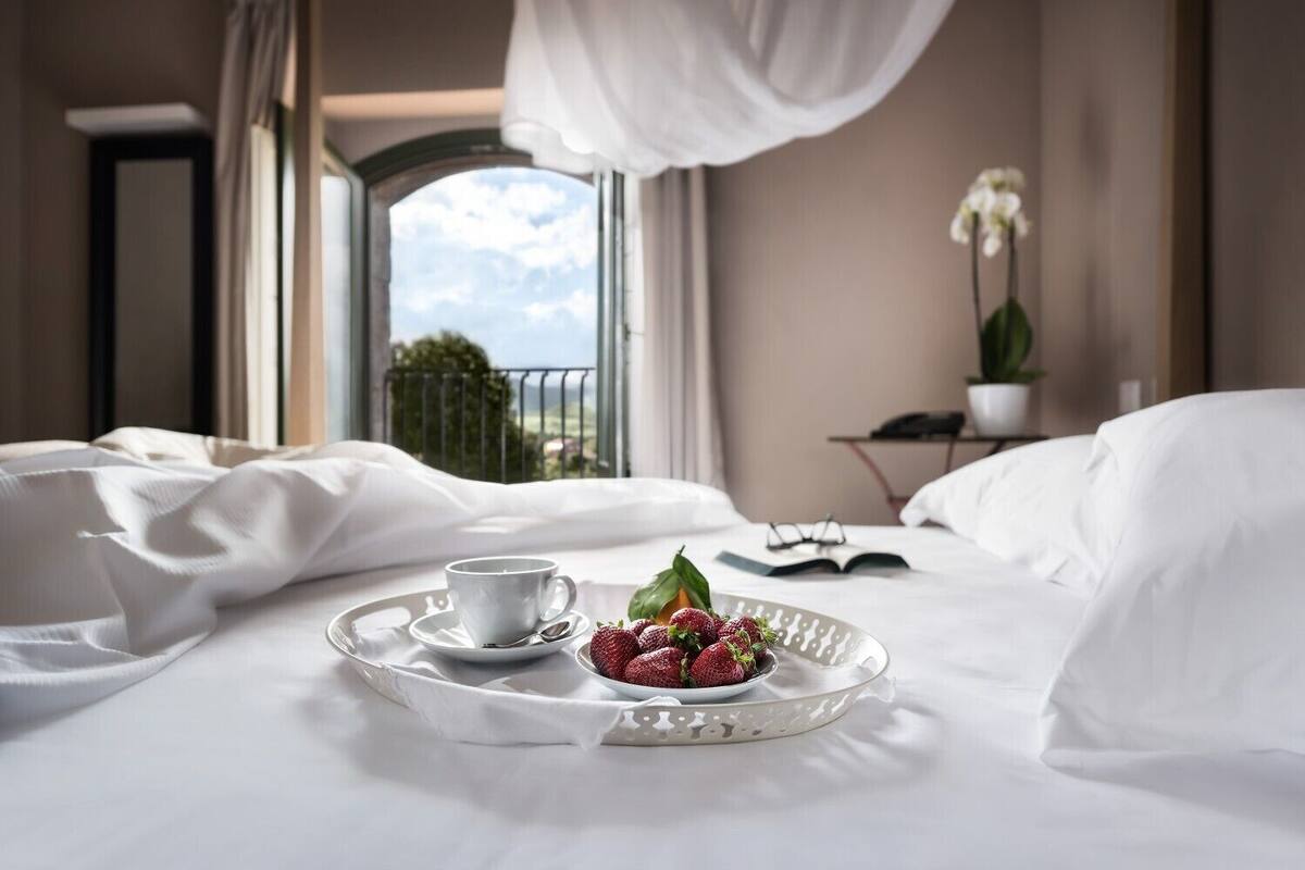 A hotel room with a bed, a tray with strawberries and a cup, a window with a view, and a plant on a side table.