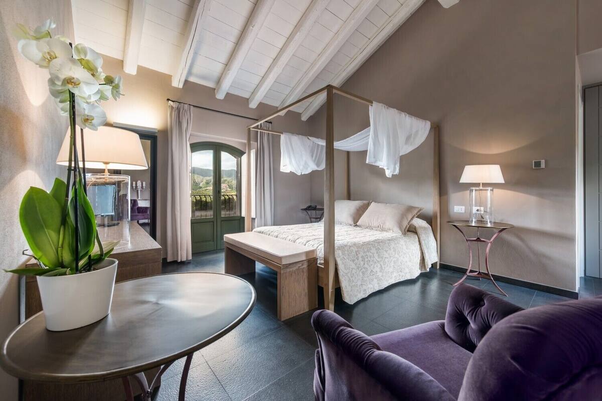 A four-poster bed with a canopy, a round table with a potted plant, a purple armchair, and a small side table with a lamp.