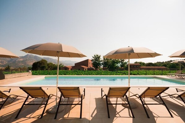 A poolside area with lounge chairs and umbrellas, a clear view of mountains, and a building in the background. - Cavanera Etnea - Resort & Wine Experience (Castiglione di Sicilia)