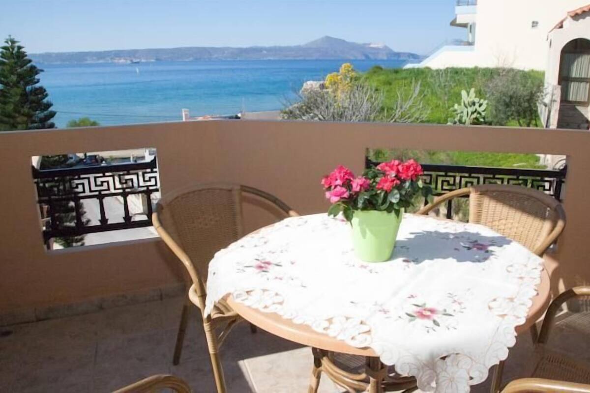 A balcony with a round table, wicker chairs, and a potted plant with pink flowers.