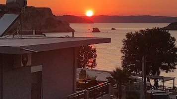 A building with a balcony overlooking a sunset over water, a parking area with cars, and a sign for 'ALOE'.