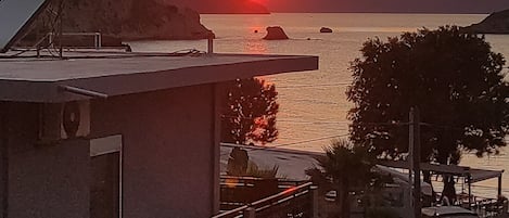 A building with a balcony overlooking a sunset over water, a parking area with cars, and a sign for 'ALOE'.