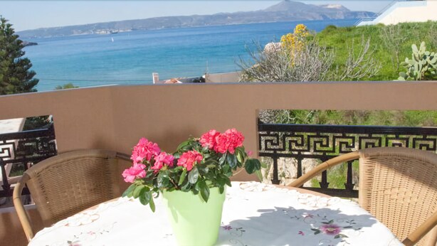 A balcony with wicker chairs, a table with a floral tablecloth, and a vase of pink flowers. - Meandros (Apokoronas)