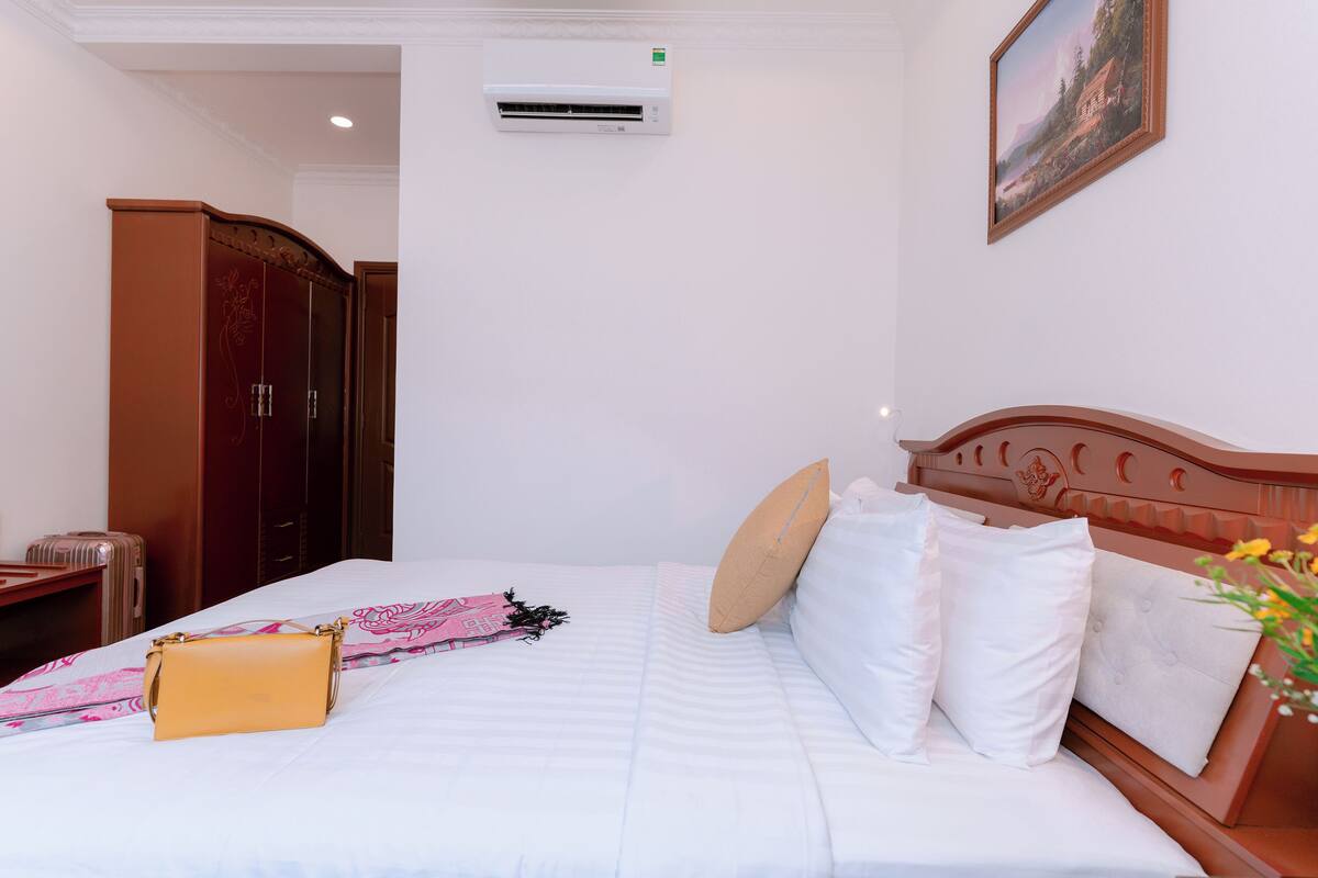 Une chambre d’hôtel avec un lit, une armoire en bois, une valise, une couverture à motifs roses et blancs, et un sac à main jaune.