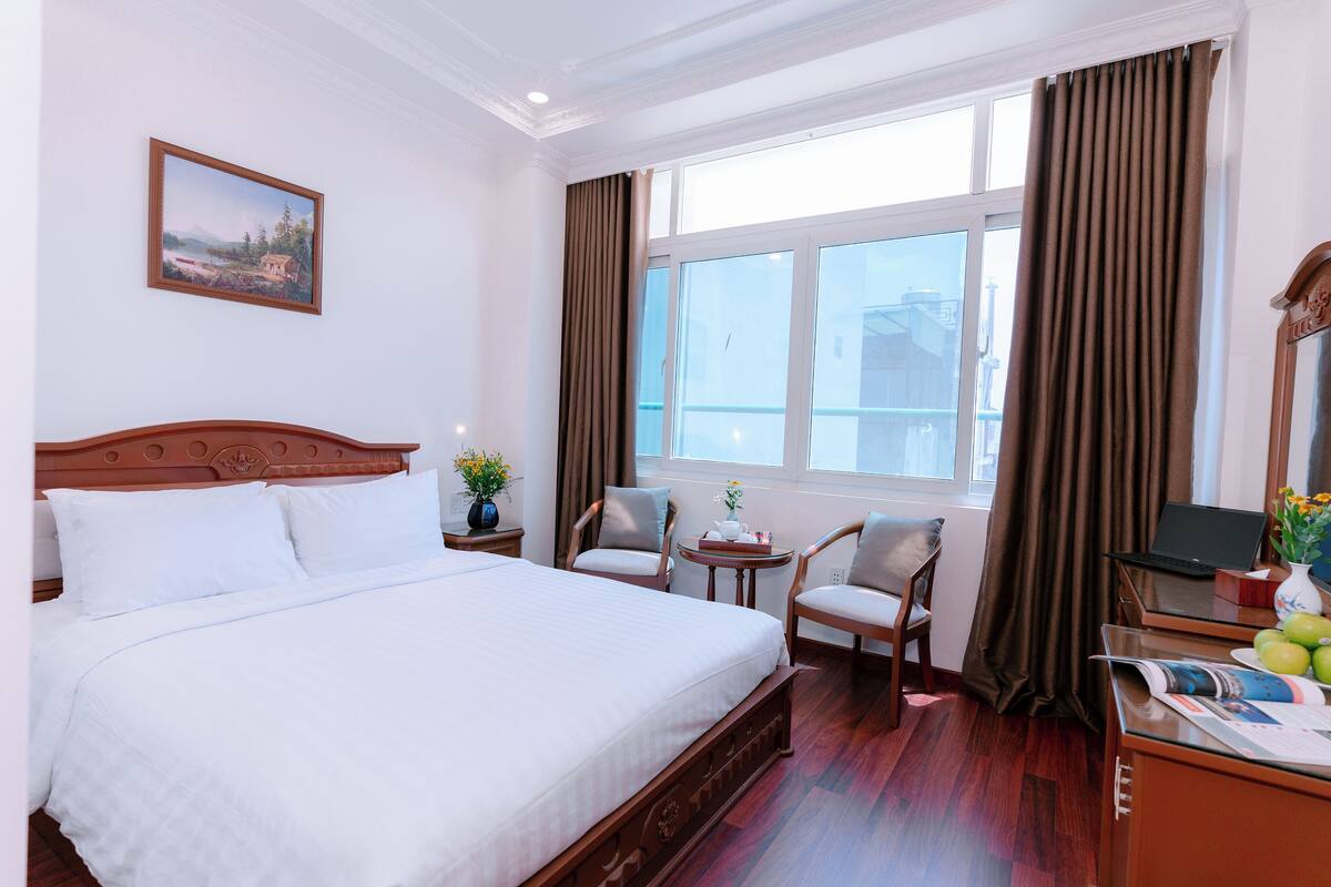Une chambre d’hôtel avec un grand lit, deux chaises, une petite table avec un vase et un ordinateur portable posé sur un bureau.