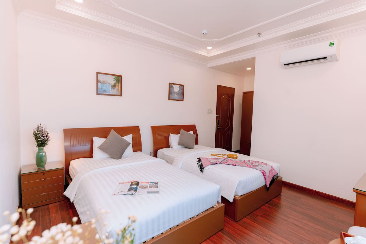 Une chambre d’hôtel avec deux lits, un plancher en bois, un vase vert avec des fleurs et un climatiseur fixé au mur.
