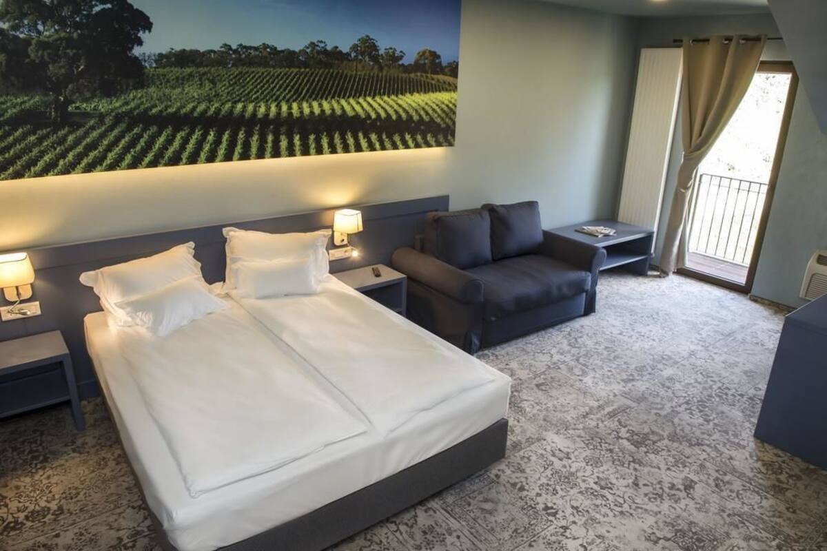 Une chambre d’hôtel avec un grand lit, un canapé, une petite table et une table de chevet. Il y a une grande fenêtre avec des rideaux et une vue sur un vignoble.