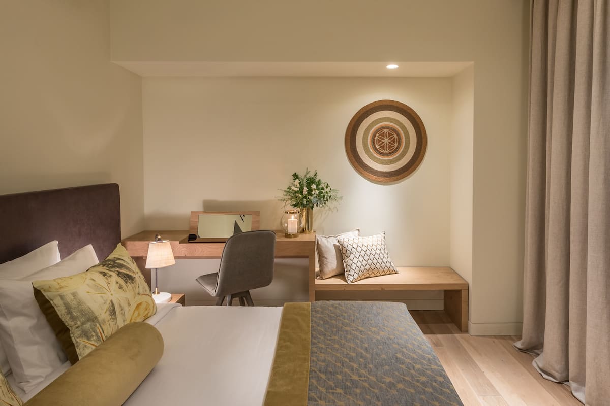 Une chambre d’hôtel comprenant un lit, un bureau, une chaise, une petite table, un élément décoratif mural et une fenêtre avec des rideaux.